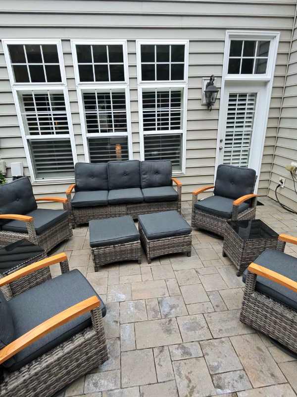 Assembled woven patio sofa and chairs with dark cushions, ottomans and side tables on a paved patio.